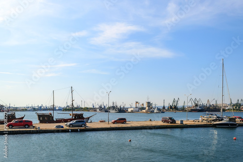 View of the water area of the small seaport with yachts, cargo ships and cranes on a clear and calm sunny day. Industrial seascape. © Maryia