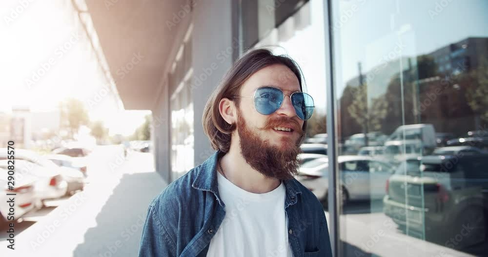 Young stylish bearded male in fancy sunglasses walking along building, smiling and feeling happy Young stylish bearded male in fancy sunglasses walking along building, smiling and feeling happy