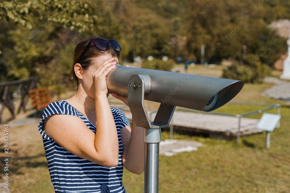 Young woman tourist in summer dress using monocular telescope binoculars to watch the scene from the mountain at the Aristotle's park at Stagira Greece in autumn