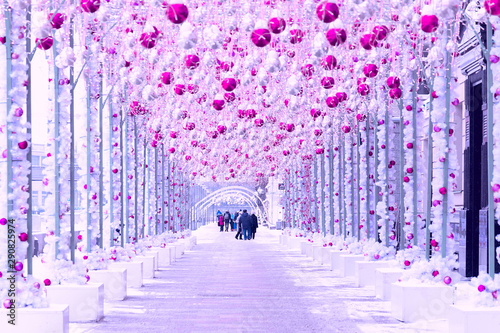 A snow-covered Christmas alley decorated with bright balls, tinsel and flickering garlands of lights along which people walk.