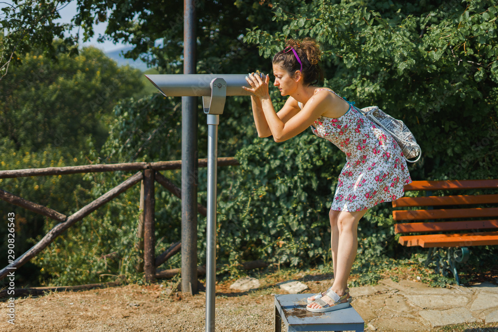 Obraz premium Young woman tourist in summer dress using monocular telescope binoculars to watch the scene from the mountain at the Aristotle's park at Stagira Greece in autumn