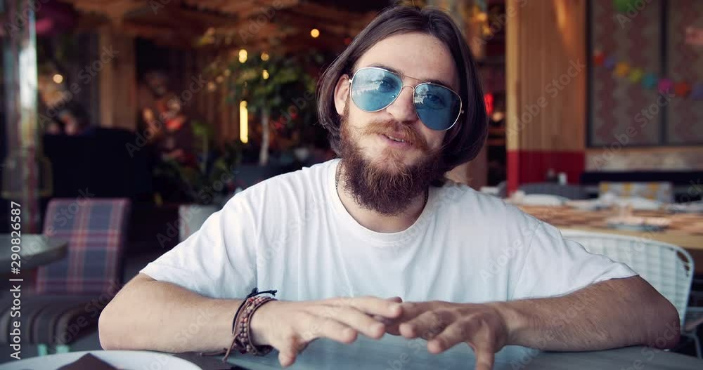 Young beaded hippie blogger in trendy eyewear recording video in fancy restaurant, sharing experience with community
