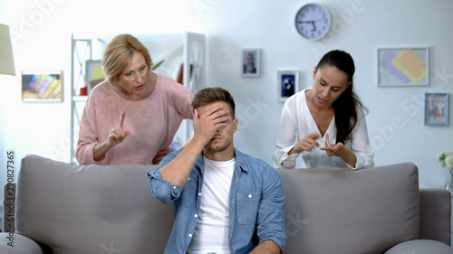 Tired man sitting on sofa, enduring to wife and mother criticize him, quarrel