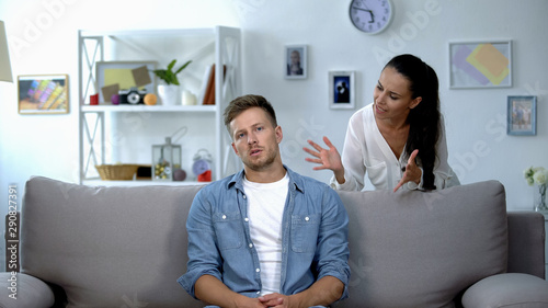 Phlegmatic man sitting on sofa listening nervous wife shouting, aggression photo