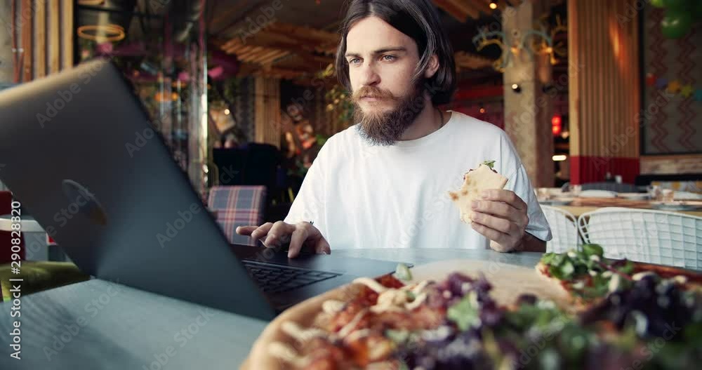 Bearded male hippie surfing internet on laptop and eating appetizing pizza in restaurant, freelancer Bearded male hippie surfing internet on laptop and eating appetizing pizza in restaurant, freelancer