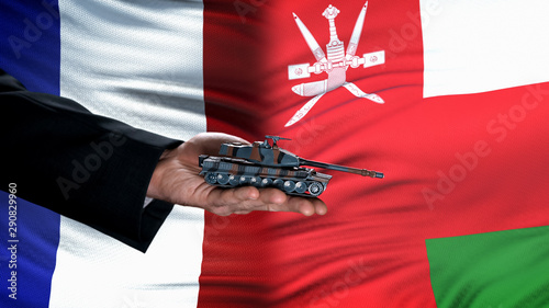 Official hand holding toy tank against France and Oman flag background, army