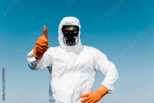 exterminator in protective mask and uniform standing with hand on hip and showing thumb up