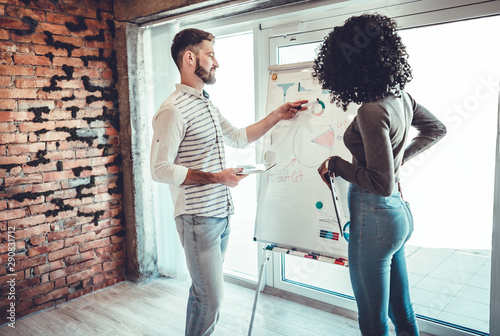 The guy and the girl are standing near the white marker board and write something on it during the working time in their office photo