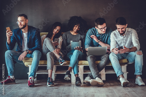 Group of happy young people sitting on sofa and using digital tablet and laptop