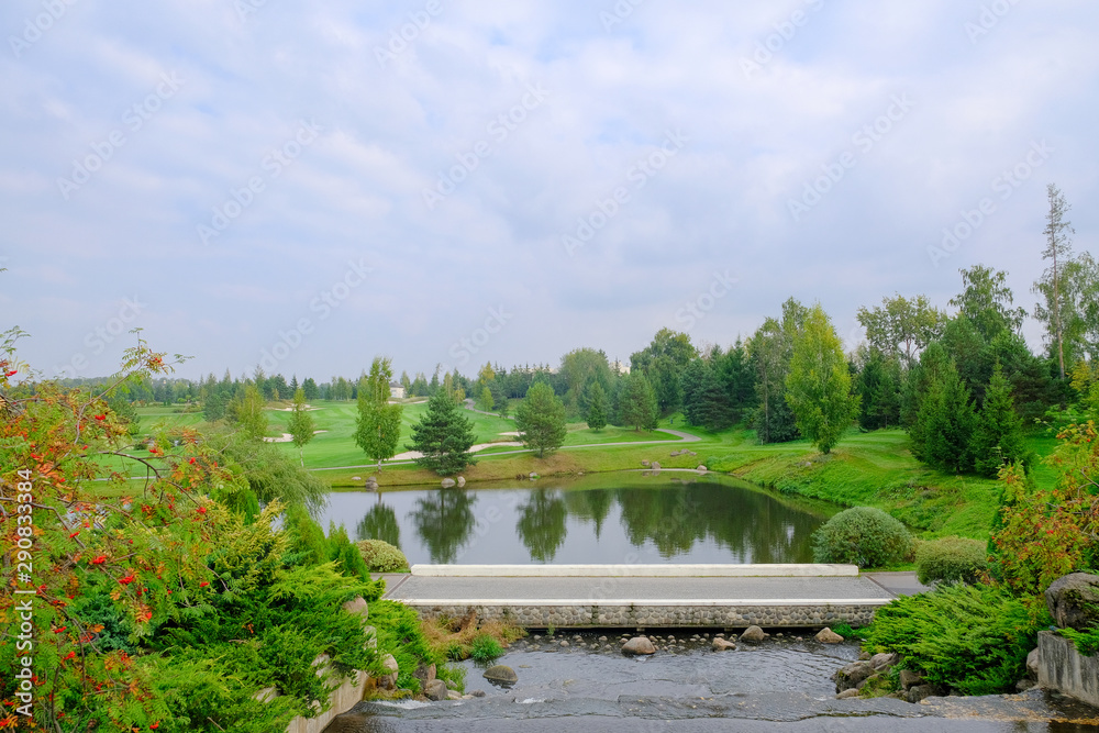 Landscape. Hills with green lawn and ornamental shrubs and trees lake and bridge. Beautiful garden