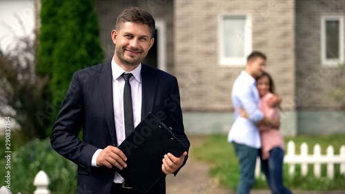 Smiling broker standing with documents, happy family hugging near their new home photo