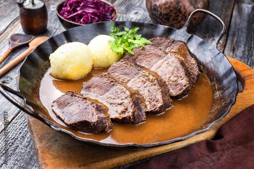 Traditioneller geschmorter deutscher Sauerbraten vom Rind mit Kartoffel Knödel und Blaukraut in würziger brauner Sauce als closeup in einer rustikalen schmiedeeisernen Pfanne photo