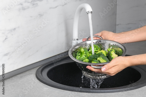 Woman washing fresh green broccoli in metal colander under tap water, closeup view. Space for text photo