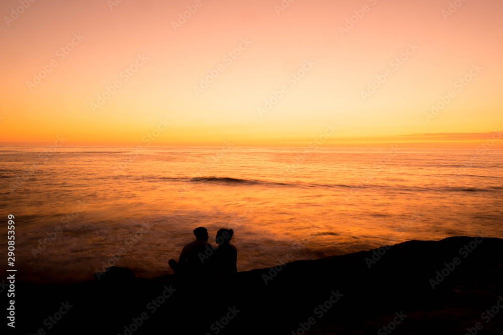 silhouette of a couple sitting on a cliff during sunset by the ocean
