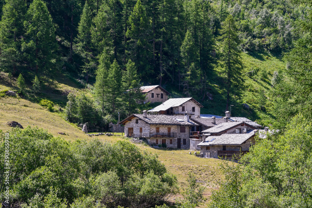 View of a small Alpine village on the slope of a mountain with typical stone houses surrounded by meadows and wood in summer, Val Ferret, Courmayeur, Val Ferret, Aosta Valley, Italy View of a small Alpine village on the slope of a mountain with typical stone houses surrounded by meadows and wood in summer, Val Ferret, Courmayeur, Val Ferret, Aosta Valley, Italy