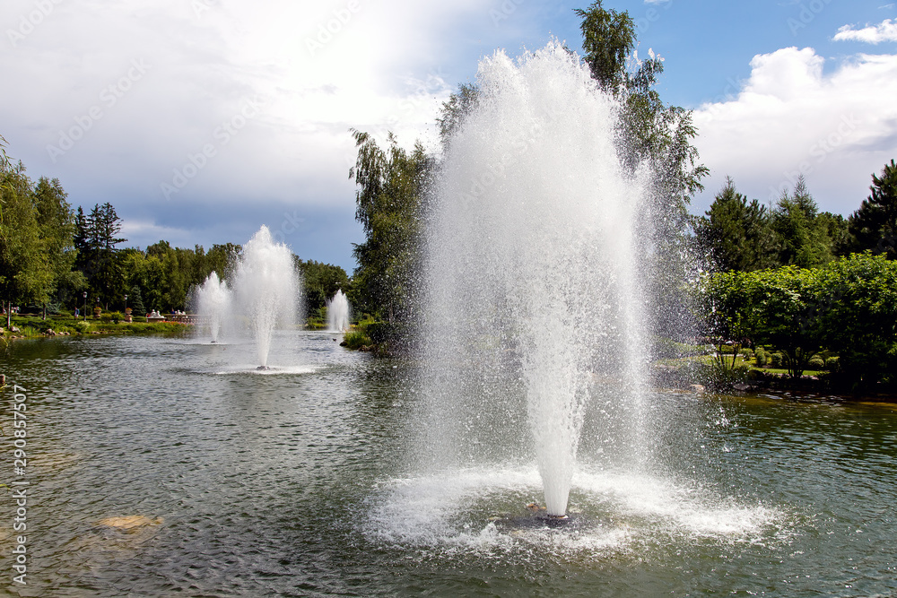 Pond with water jets of a fountain in a summer park lit by sunlight with a cloudy sky.