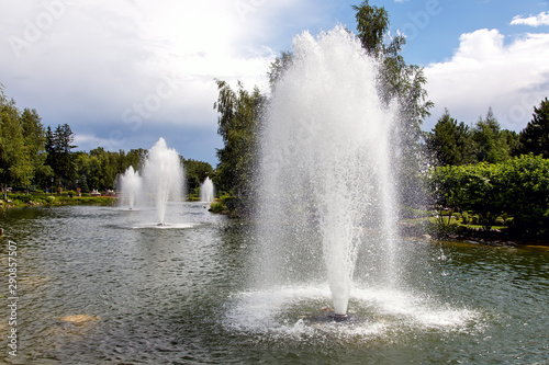 Pond with water jets of a fountain in a summer park lit by sunlight with a cloudy sky.