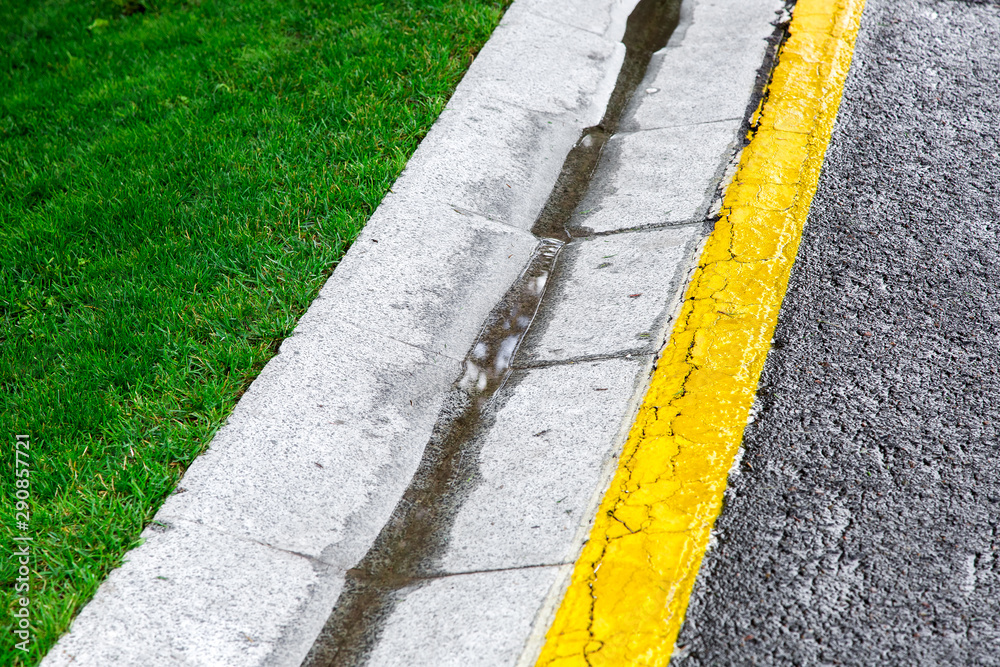 flowing rainwater in the canal is a cement ditch of a drainage system on the side of an asphalt road with yellow markings and a green lawn.