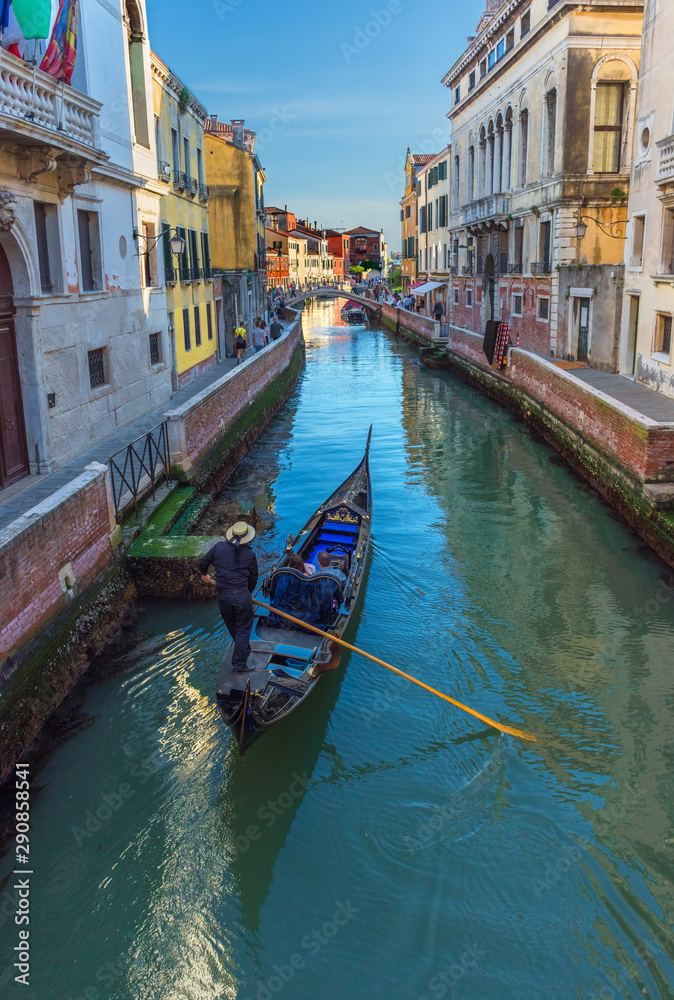  Venetian canal with tourists in the gondola among boats and historic houses, in a beautiful sunny day.