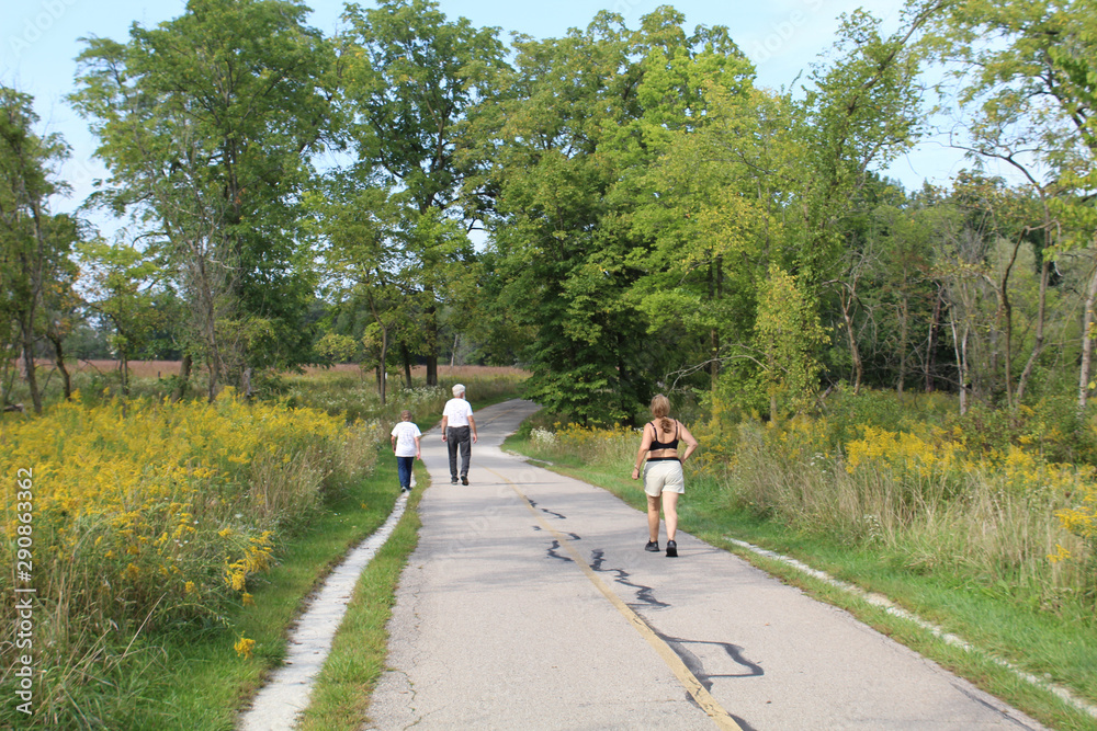 Older couple walking with a middle-aged woman running behind them on the North Branch Trail with goldenrod at Miami Woods in Morton Grove, Illinois
