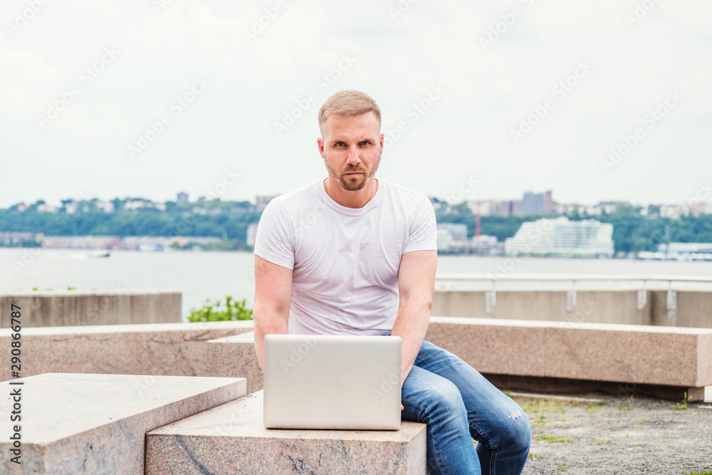 Young American Man with beard, reading, working on laptop computer outside in New York City, wearing white T shirt, blue jeans, sitting on stone bench at park, reading, working on laptop computer,.