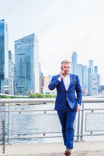 Young American Businessman with beard, talking on cell phone, traveling in New York City, wearing blue suit, white shirt, walking in business district with high buildings in Midtown of Manhattan.. © Alexander Image