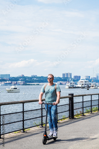 Fototapeta Naklejka Na Ścianę i Meble -  Young American Man traveling in New York City, wearing light green T shirt, blue jeans, sneakers, sunglasses, shoulder carrying back bag, riding on electric scooter, pass by Hudson River with boats..