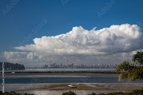 Auckland city scape from West Harbour. Looking out to sky tower and surrounding buildings across the harbour and marshes, wetlands. Water is still, low tide. Clouds are building over waitemata harbour
