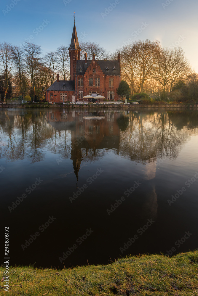 Heritage of brick building reflects into lake with dry tree background at Minnewater park during sunrise, Bruges, Belgium