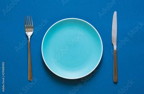 Ready to order. High angle shot of empty color plate, fork, knife lying against blue background.