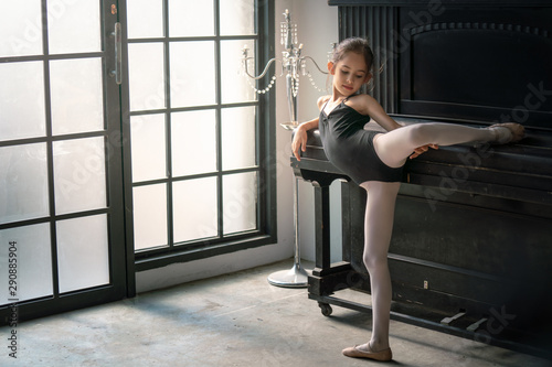 Pretty little child girl ballet dancer in black leotard and ballet slipper shoes do dance practice at vintage style studio room with piano. Happy young schoolgirl ballerina dancing ballet in the room.