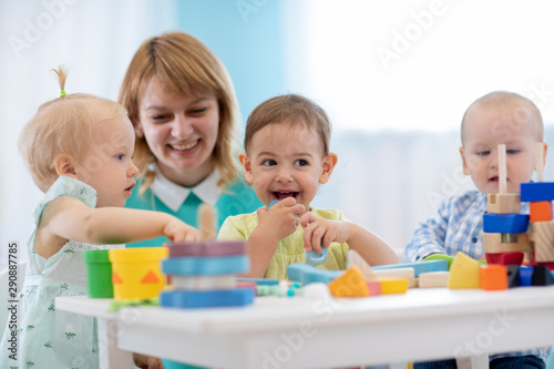 Children group playing with teacher in day care centre. Carer and babies sitting at table in nursery playroom