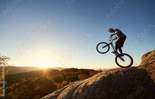 Silhouette of cyclist balancing on back wheel on trial bicycle. Courageous sportsman biker making acrobatic trick on top of rocky mountain at sunset. Concept of extreme sport active lifestyle