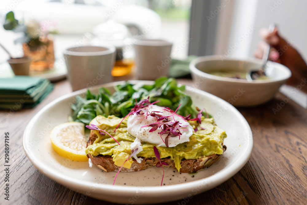 Morning in cafe, oak table. Healthy breakfast with wholemeal bread toast with avocado, poached egg with green salad. Green tea on the Background.