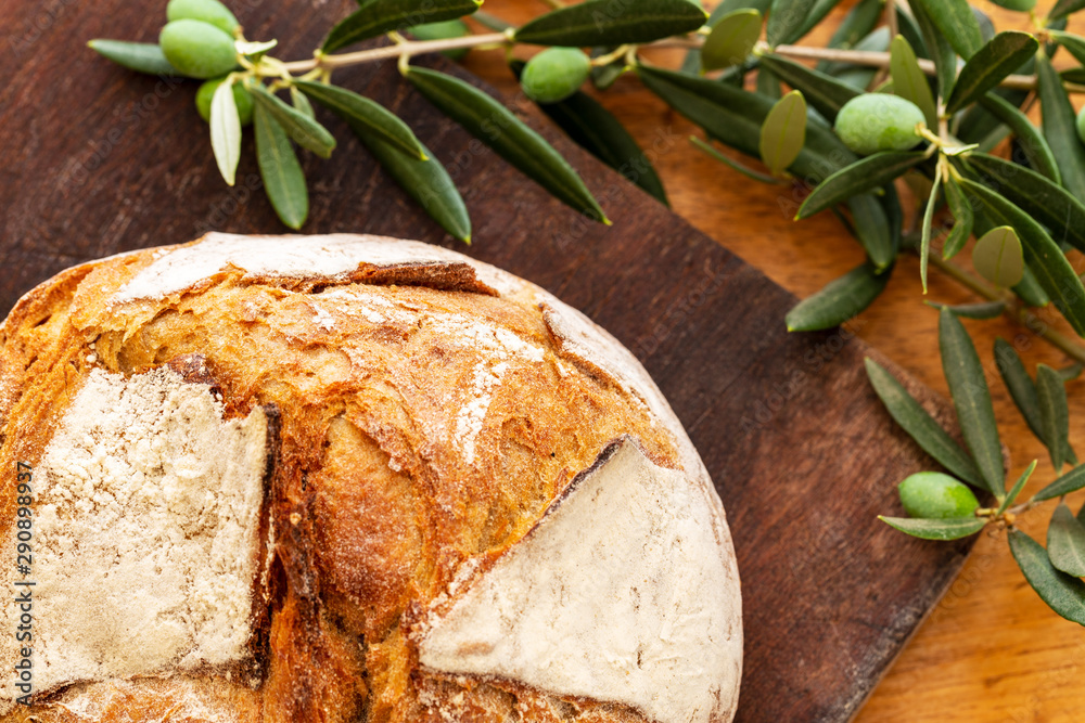 Φωτογραφία Bread loaf covered with flour, on a dark wooden board with green olives on branch or twig with leaves over a vintage used oak wood background