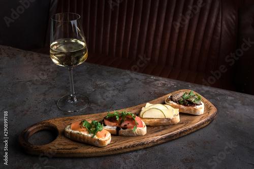 Four bruschettas lay on wooden board. Appetizers with tomato, mozzarella, pesto, salmon and roast beef. A glass of white wine on on a gray concrete table in a restaurant. place for text