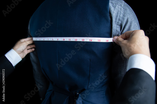 The dressmaker was measuring the width of the back of the man wearing a blue suit on black background.