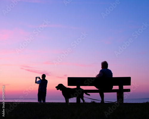 man takes pictures of colorful sunset while woman sits with dog on bench © ahavelaar