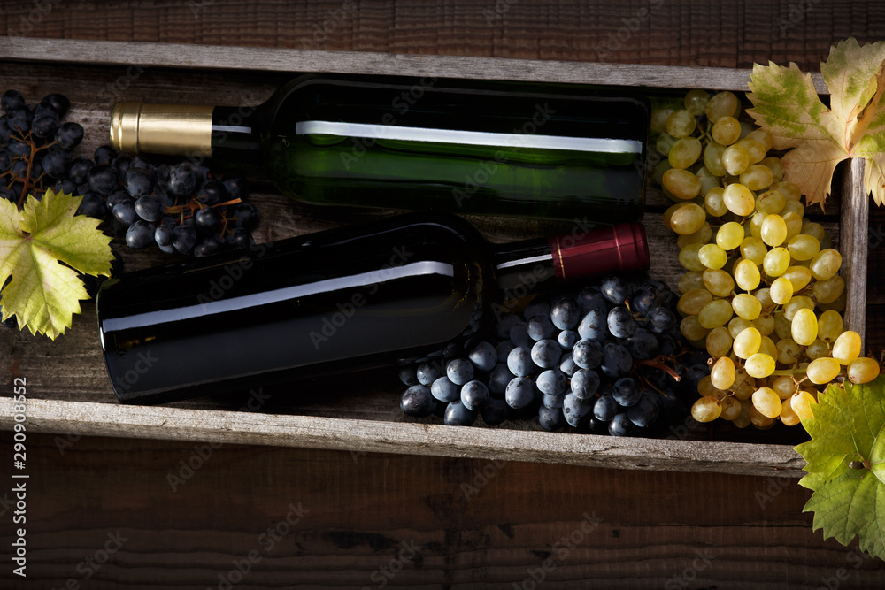Obraz premium A bottle of red wine and a bottle of white wine, black grapes and white grapes with grape leaves on an old wooden table. Dark background.