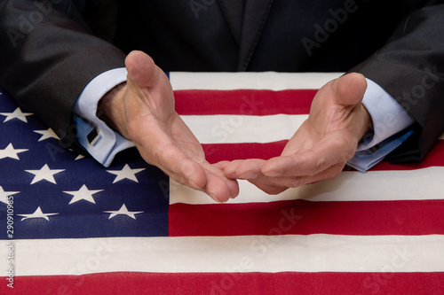 At a table covered with an American flag, a male businessman and politician holds out his hands for dialogue and suggestions