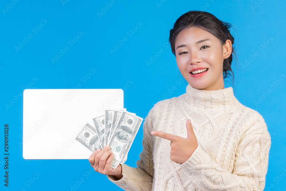 A beautiful woman wearing a new long-sleeved white carpet holding a white sign and a dollar bill on a blue background. Trading concept. A beautiful woman wearing a new long-sleeved white carpet holding a white sign and a dollar bill on a blue background. Trading concept.
