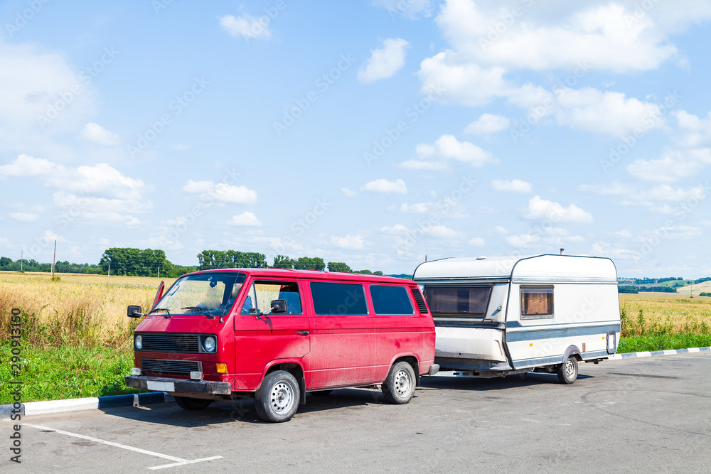 A motor home in the form of a white trailer in a parking lot near the road, fastened to a red car in the back of a minivan while traveling on a summer day against a blue sky. Movable property.