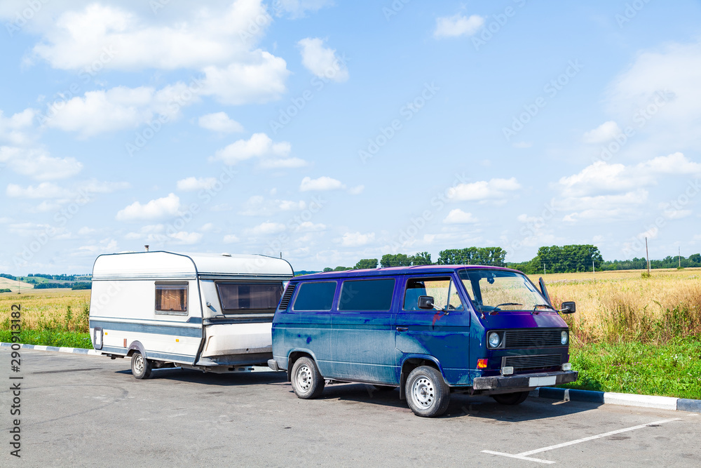 A motor home in the form of a white trailer in a parking lot near the road, fastened to a blue car in the back of a minivan while traveling on a summer day against a blue sky. Movable property.