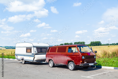 A motor home in the form of a white trailer in a parking lot near the road, fastened to a red car in the back of a minivan while traveling on a summer day against a blue sky. Movable property.