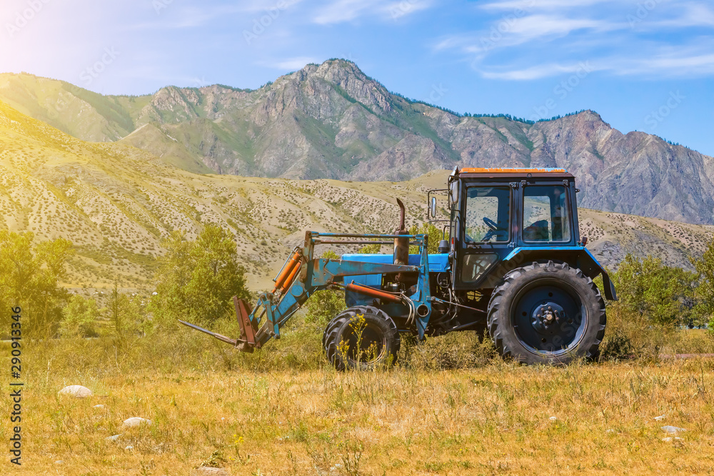 An old blue tractor excavator stands in a field with green and yellow grass against a background of mountains and blue sky during harvesting in summer or autumn. Agriculture and farming machinery. An old blue tractor excavator stands in a field with green and yellow grass against a background of mountains and blue sky during harvesting in summer or autumn. Agriculture and farming machinery.