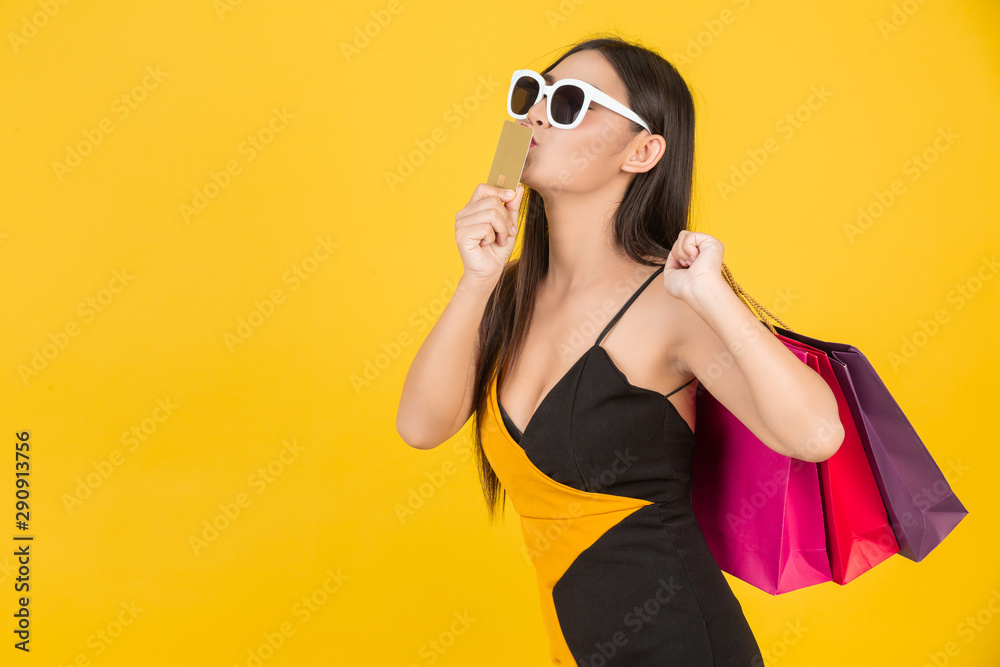 Shopping concept of a beautiful woman wearing glasses with a gold credit card with a colorful paper bag on a yellow background. Shopping concept of a beautiful woman wearing glasses with a gold credit card with a colorful paper bag on a yellow background.