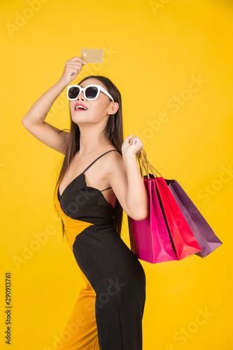Shopping concept of a beautiful woman wearing glasses with a gold credit card with a colorful paper bag on a yellow background.