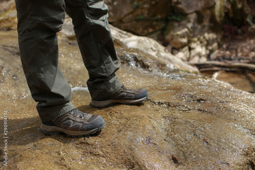 Close up view of boots in water. Person in green trousers and in brown trekking shoes stands on a stone through which a mountain stream flows. Close up view of boots in water. Person in green trousers and in brown trekking shoes stands on a stone through which a mountain stream flows.