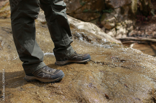 Close up view of boots in water. Person in green trousers and in brown trekking shoes stands on a stone through which a mountain stream flows.