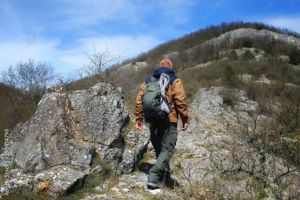 A red-haired man in a brown windbreaker with backpack and rope walks past a stone on a mountain slope in sunny weather. There are no foliage on trees and bushes.